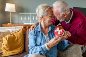 senior couple sitting on a couch in their living room smiling at each other touching foreheads while woman holds a heart-shaped gift